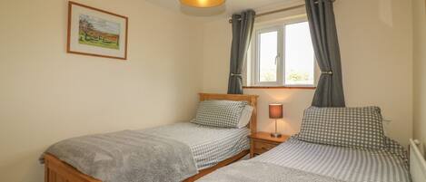 A bedroom with two single beds, a bedside table with a lamp, a window with curtains, and a framed picture on the wall.