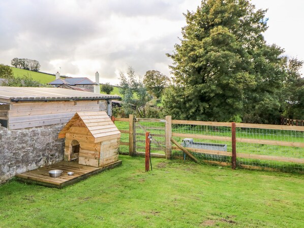 A rustic wooden structure with a sloped roof, a small wooden platform with a metal bowl, and a fenced area with a wire gate. - Higher Whiddon Farm Whiddon Well (Newton Abbot)