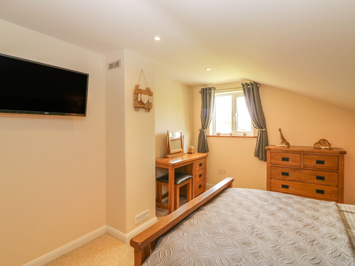 A bedroom with a bed, a dresser, a vanity, a television, and a window with curtains.