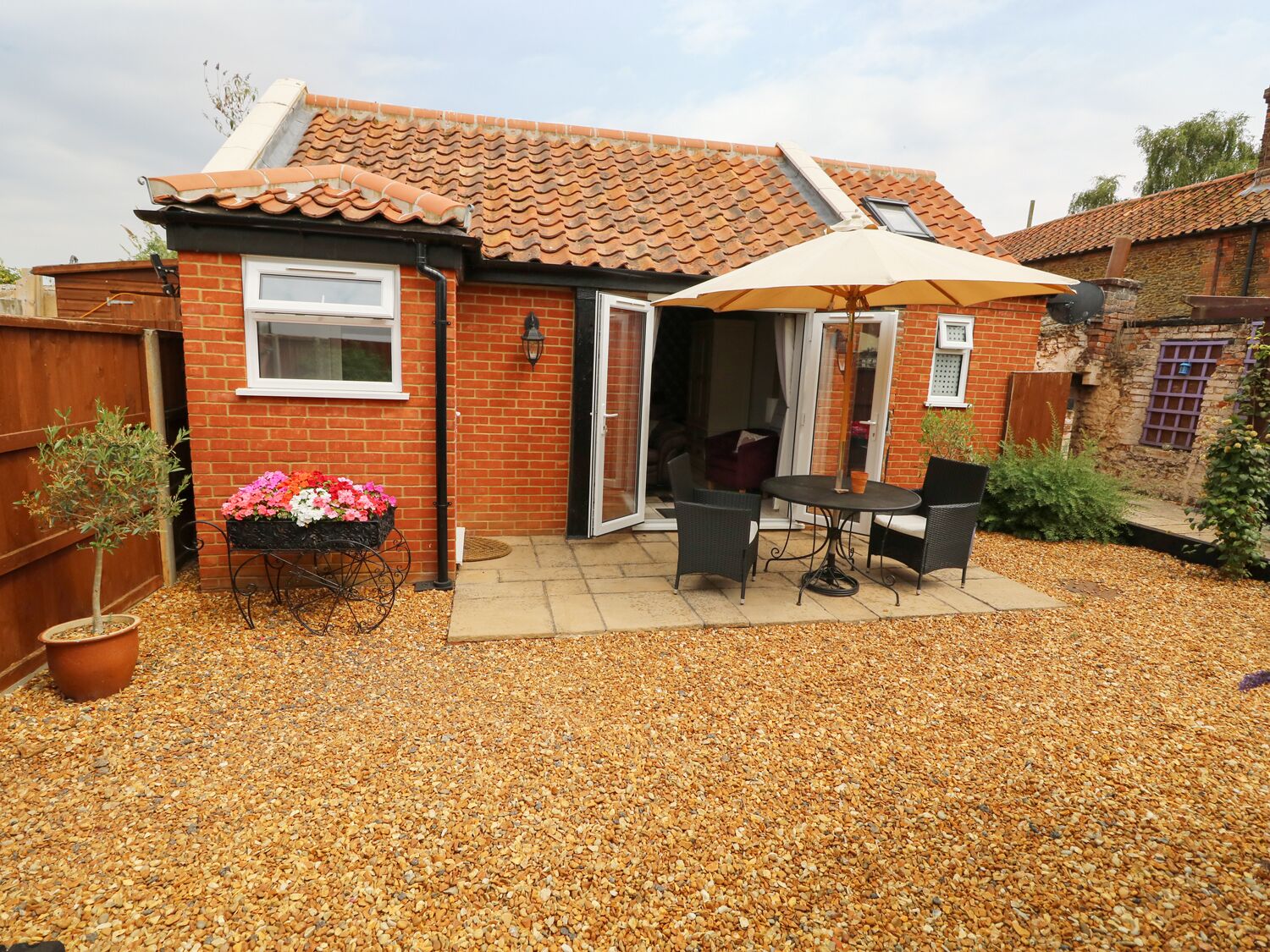 A brick house with a tiled roof, a patio with a table and chairs, and a flower bed with pink and white flowers.