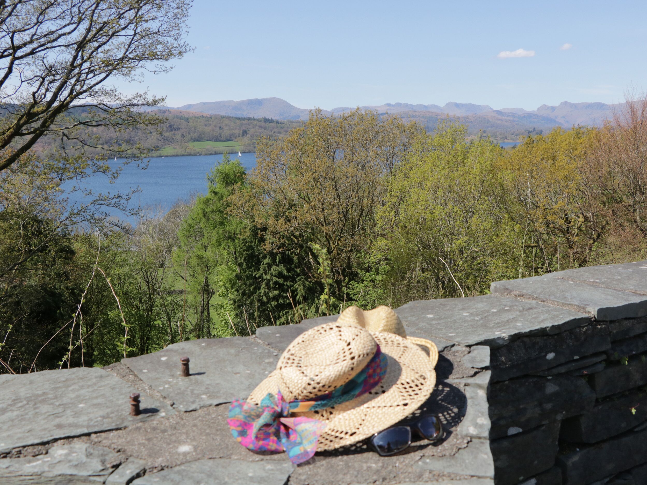 A stone wall with a straw hat and sunglasses placed on it, overlooking a lake and forested hills.