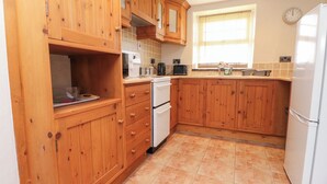 A kitchen with wooden cabinets, a white refrigerator, a microwave, and a window with blinds. - Garreg Wen Isaf Bach (Caernarfon)