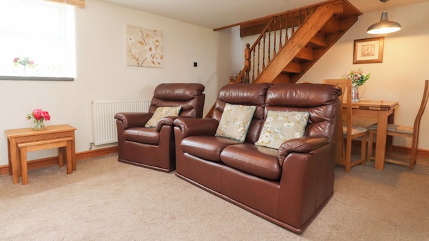A living room with two brown leather sofas, a wooden table, a wooden chair, a small wooden side table with a vase of flowers, a radiator, a staircase, and a framed picture on the wall. - Garreg Wen Isaf Bach (Caernarfon)