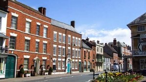 A street view with traditional brick buildings, a clear blue sky, and a flower bed with various flowers. - Cider Barn Cottage (Tewkesbury)