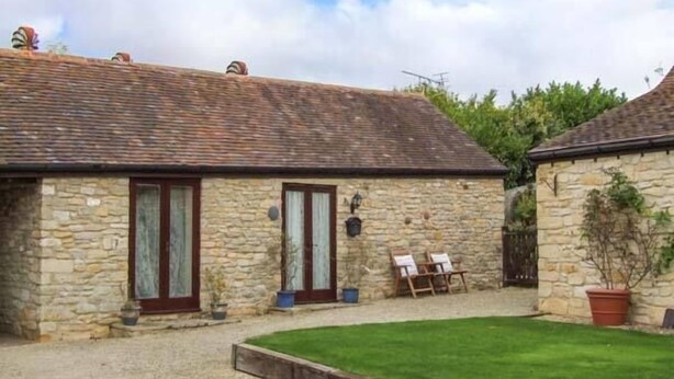 A stone building with a tiled roof, a grassy yard, and a small garden area with a chair and table. - Cider Barn Cottage (Tewkesbury)