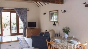 A cozy living room with a television, a sofa, a dining table set for two, and a window with curtains. - Cider Barn Cottage (Tewkesbury)