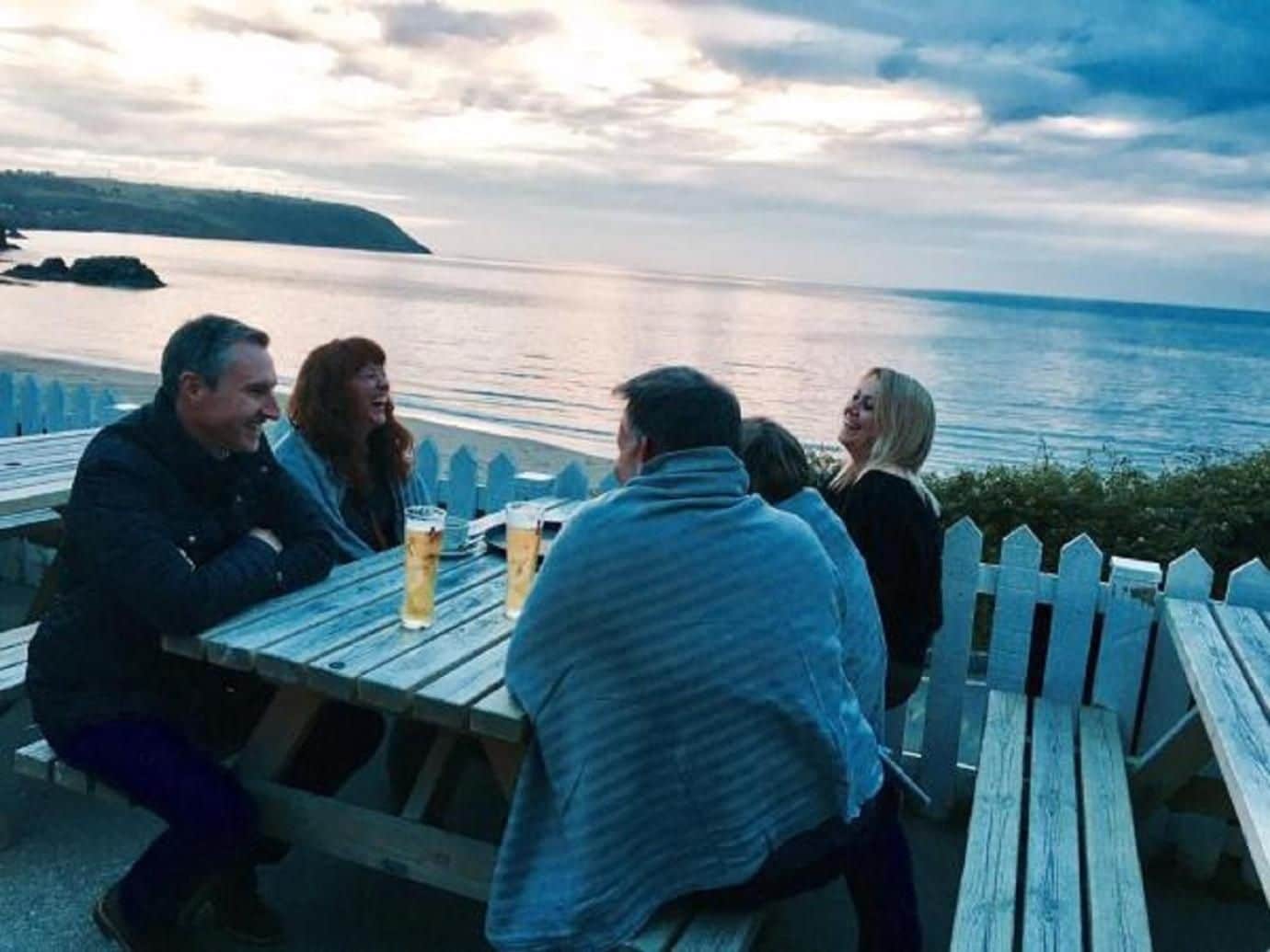 Een groep mensen zit aan een houten tafel buiten, met drankjes, met uitzicht op een wateroppervlak en een landschap in de verte.