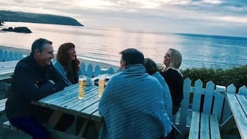 Een groep mensen zit aan een houten tafel buiten, met drankjes, met uitzicht op een wateroppervlak en een landschap in de verte.