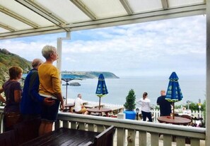 A covered outdoor seating area with tables and umbrellas, overlooking a body of water and a distant landmass.
