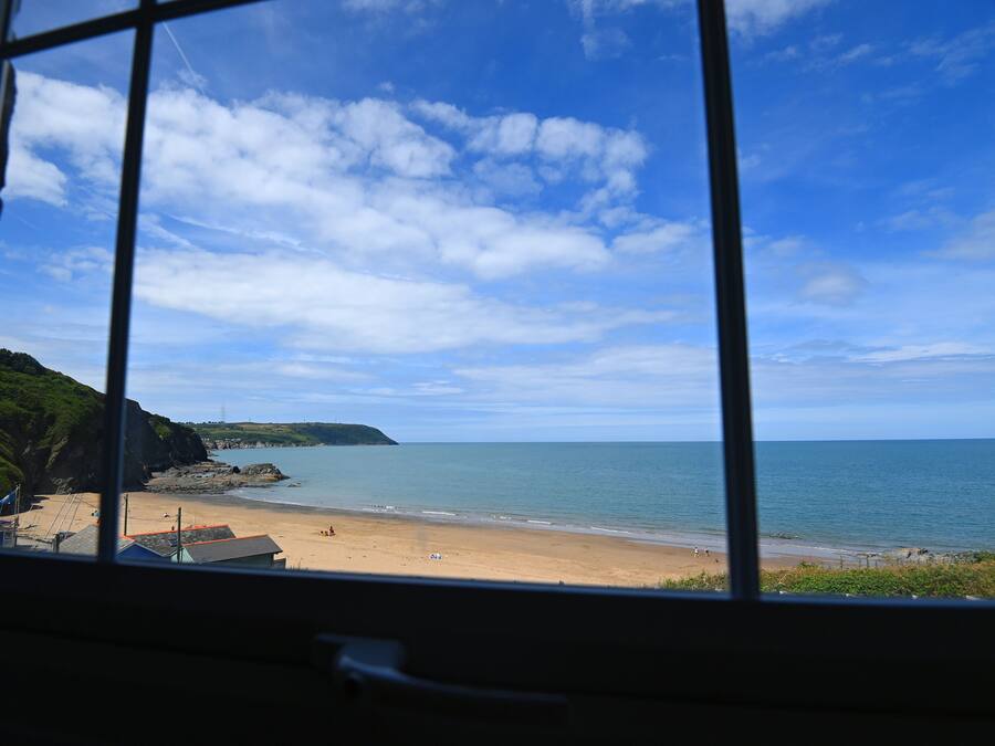 Vista costeira através de uma janela, com uma praia arenosa, mar calmo e céu claro.