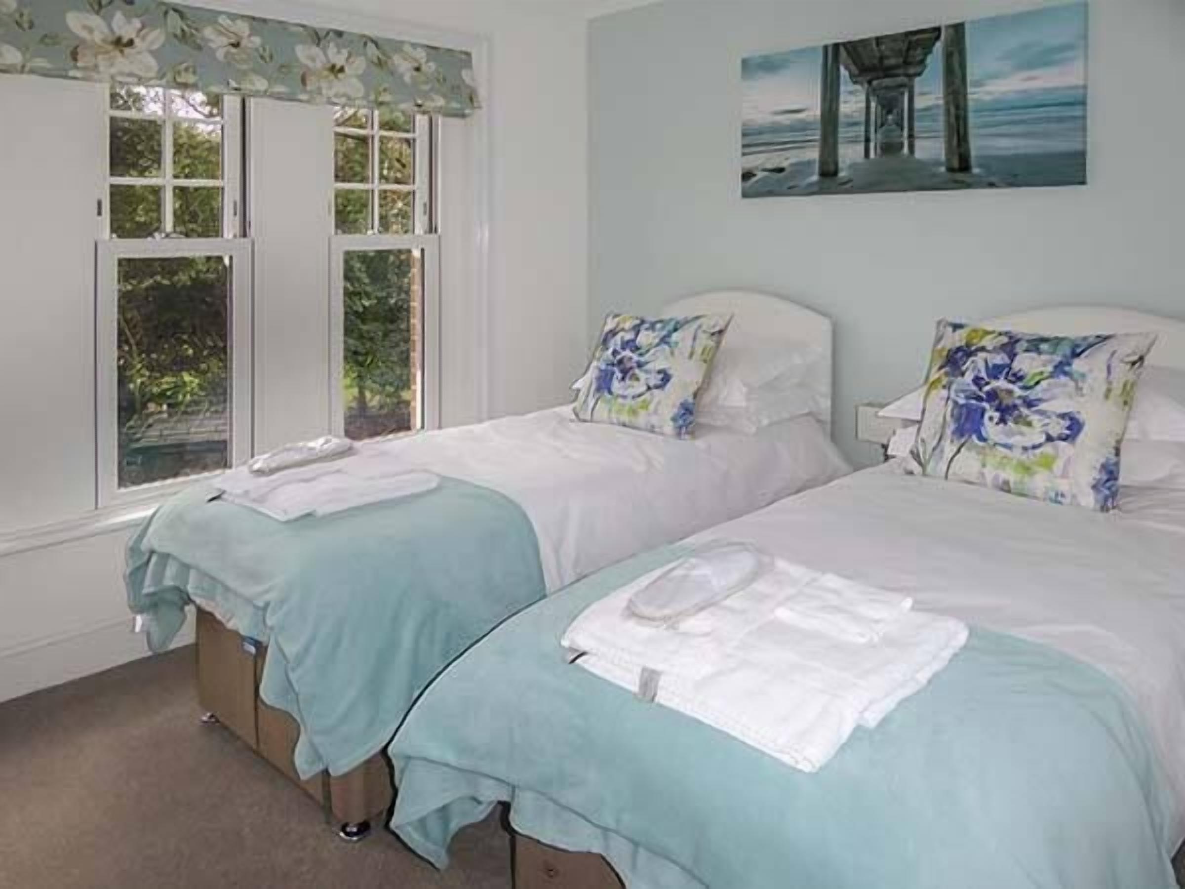 A bedroom with two beds, a window with floral curtains, and a painting of a pier on the wall.