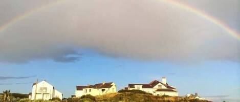 A coastal road with houses on a hillside, a stone wall, and a rainbow in the sky.