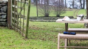 Un espace extérieur rustique aménagé avec une table et des chaises en bois, un mur en pierre et une vue sur un paysage verdoyant, avec des arbres et une clôture en arrière-plan.