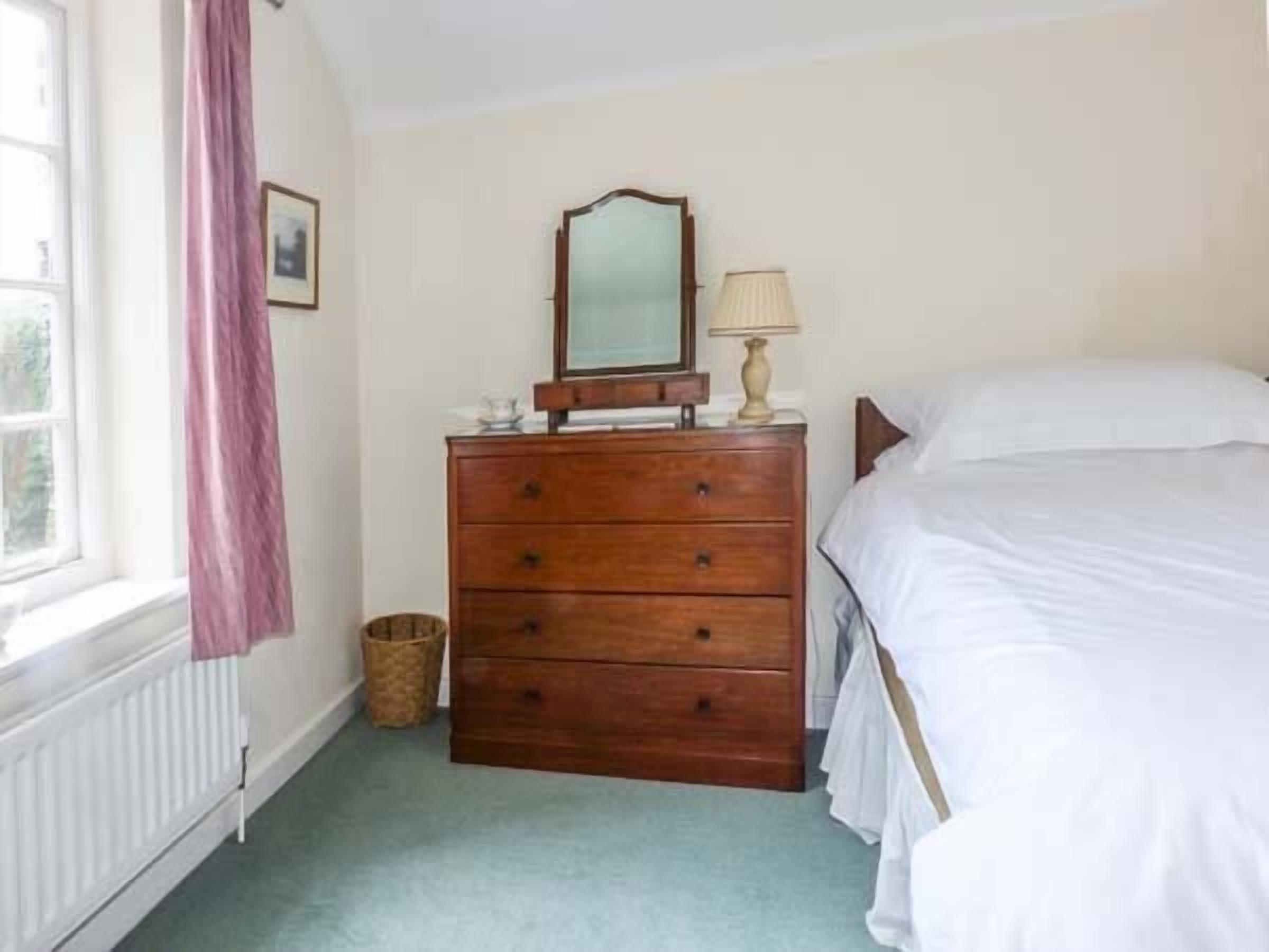 A bedroom with a wooden dresser, a bed, a window with a curtain, and a lamp on the dresser.