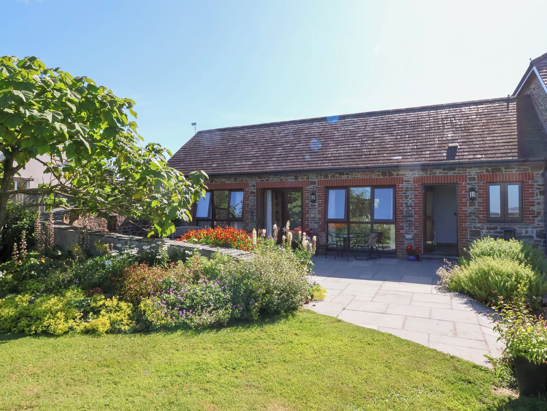 A stone building with a tiled roof, a paved patio, and a variety of flowering plants.