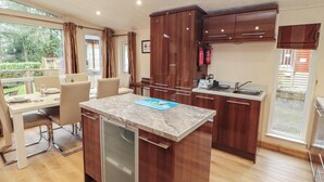 A modern kitchen with wooden cabinets, a built-in dishwasher, and a dining area with a table and chairs. - Beech Hill Lodge at Fallbarrow Park (Windermere)