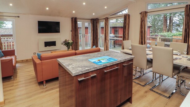 A modern kitchen with a central island, wooden cabinets, and a dining area with a table and chairs. - Beech Hill Lodge at Fallbarrow Park (Windermere)