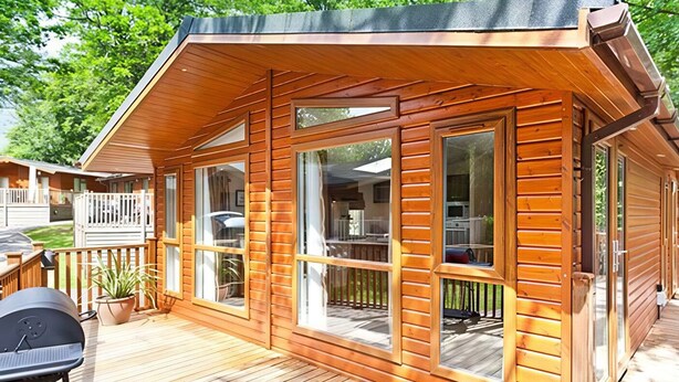 A wooden cabin with large windows, a deck, and a barbecue grill. - Beech Hill Lodge at Fallbarrow Park (Windermere)