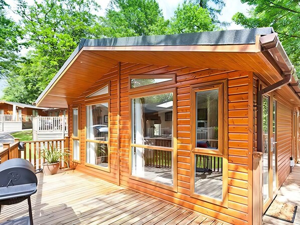A wooden cabin with large windows, a deck, and a barbecue grill. - Beech Hill Lodge at Fallbarrow Park (Windermere)