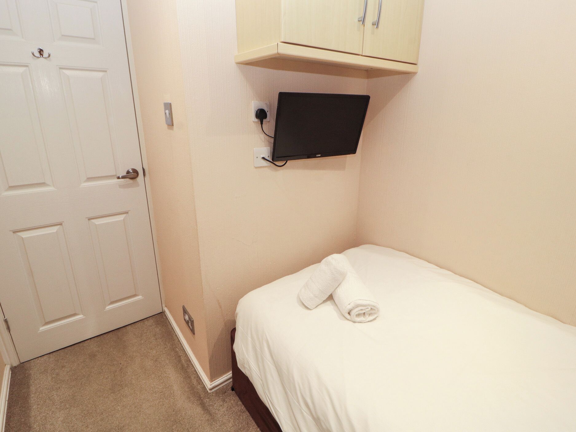 Ein kleines, ordentlich eingerichtetes Schlafzimmer mit einem Einzelbett, einem an der Wand montierten Fernseher und einer Tür.