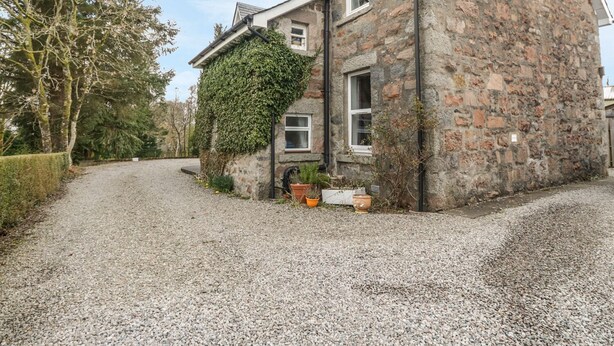 A stone house with a gravel driveway, a small garden, and a large tree to the left. - The Knowe Upper (Taynuilt)