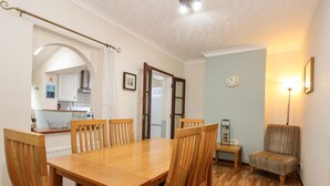 A dining area with a wooden table and chairs, a kitchen in the background, and a small armchair. - 5 Melinda Cottages (Cromer)