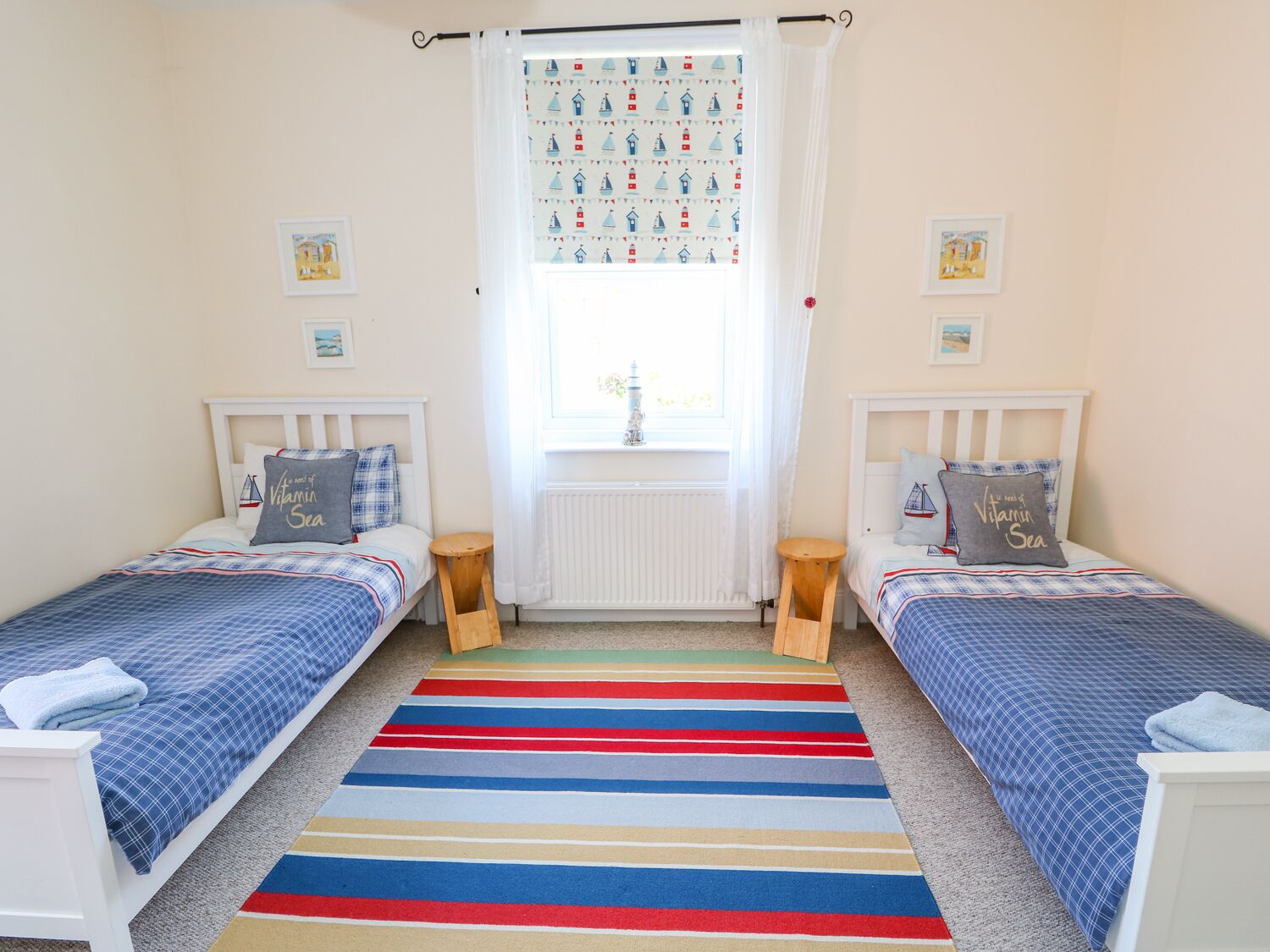 A bedroom with two single beds, a colorful striped rug, a window with a sailboat curtain, and a radiator.