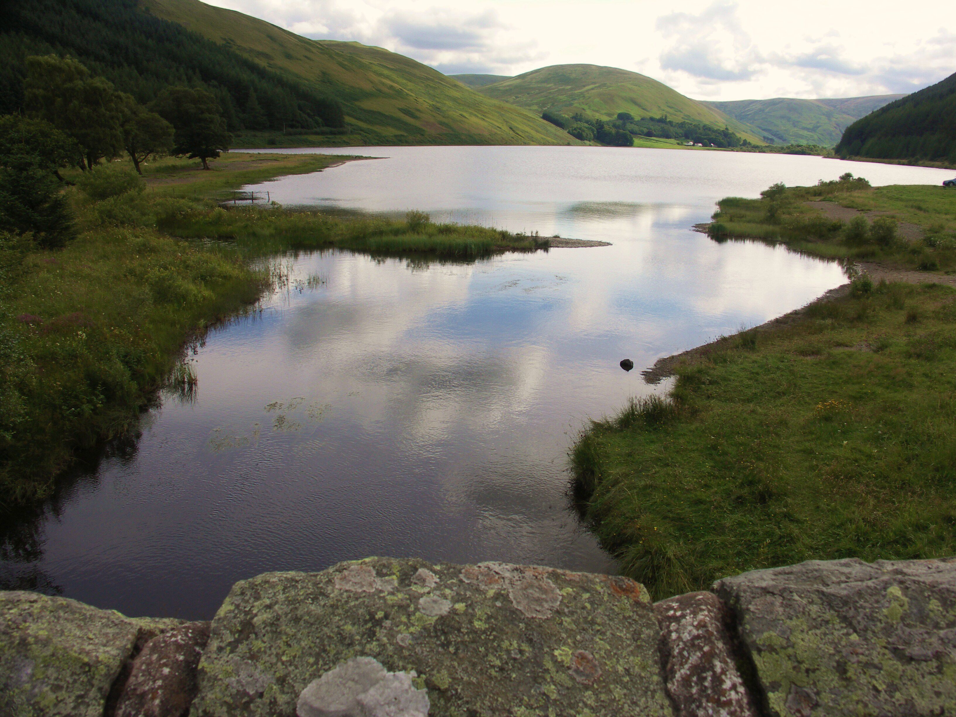 A calm lake surrounded by green hills and a stone wall in the foreground.