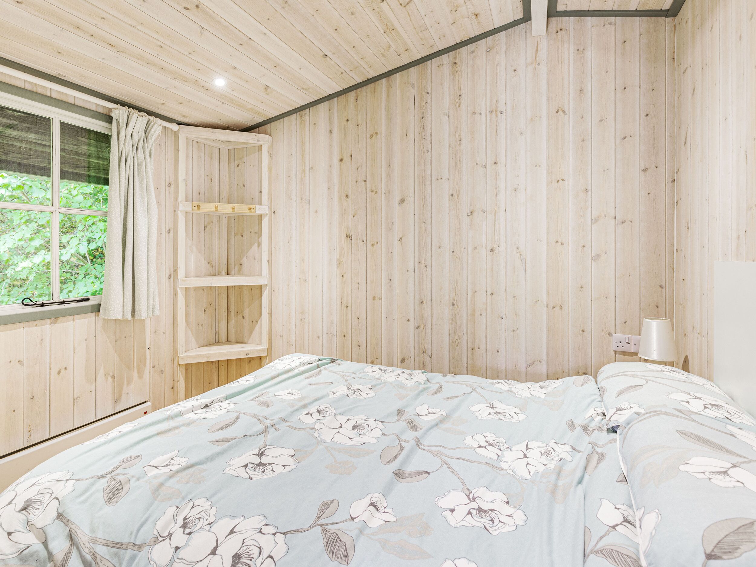 Un dormitorio con paredes de madera, una cama con ropa de cama floral, una ventana con cortinas y una estantería.