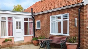 A brick house with a tiled roof, a white door, and a window with a curtain. There is a bench and a table outside. - Glenhaven (Saxmundham)