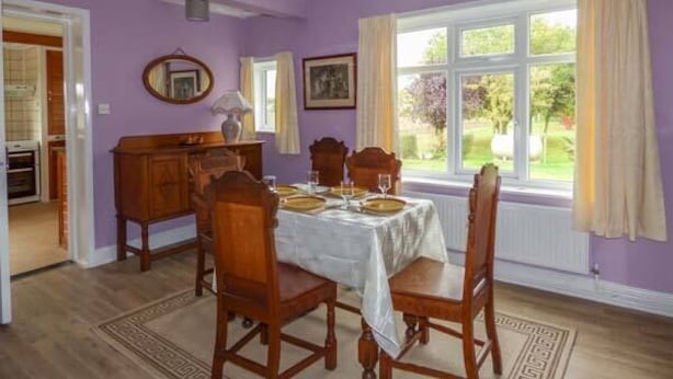 A dining room with a table set for four, a wooden cabinet, and a window with curtains. - Glenhaven (Saxmundham)