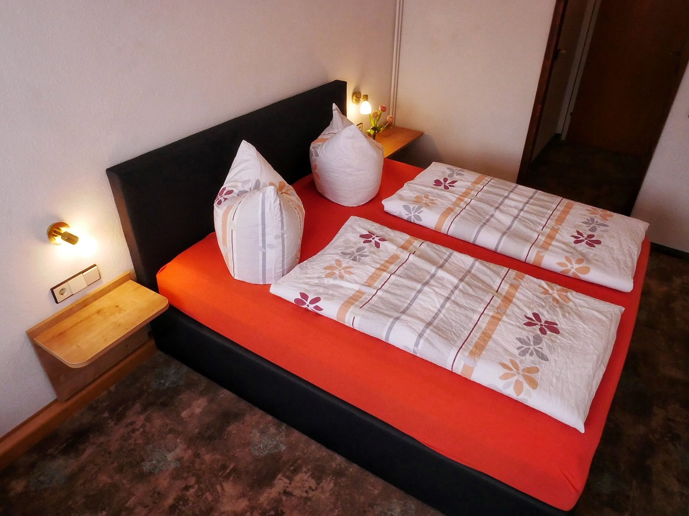 A bed with a red mattress, two pillows, and a bedside table with a lamp.