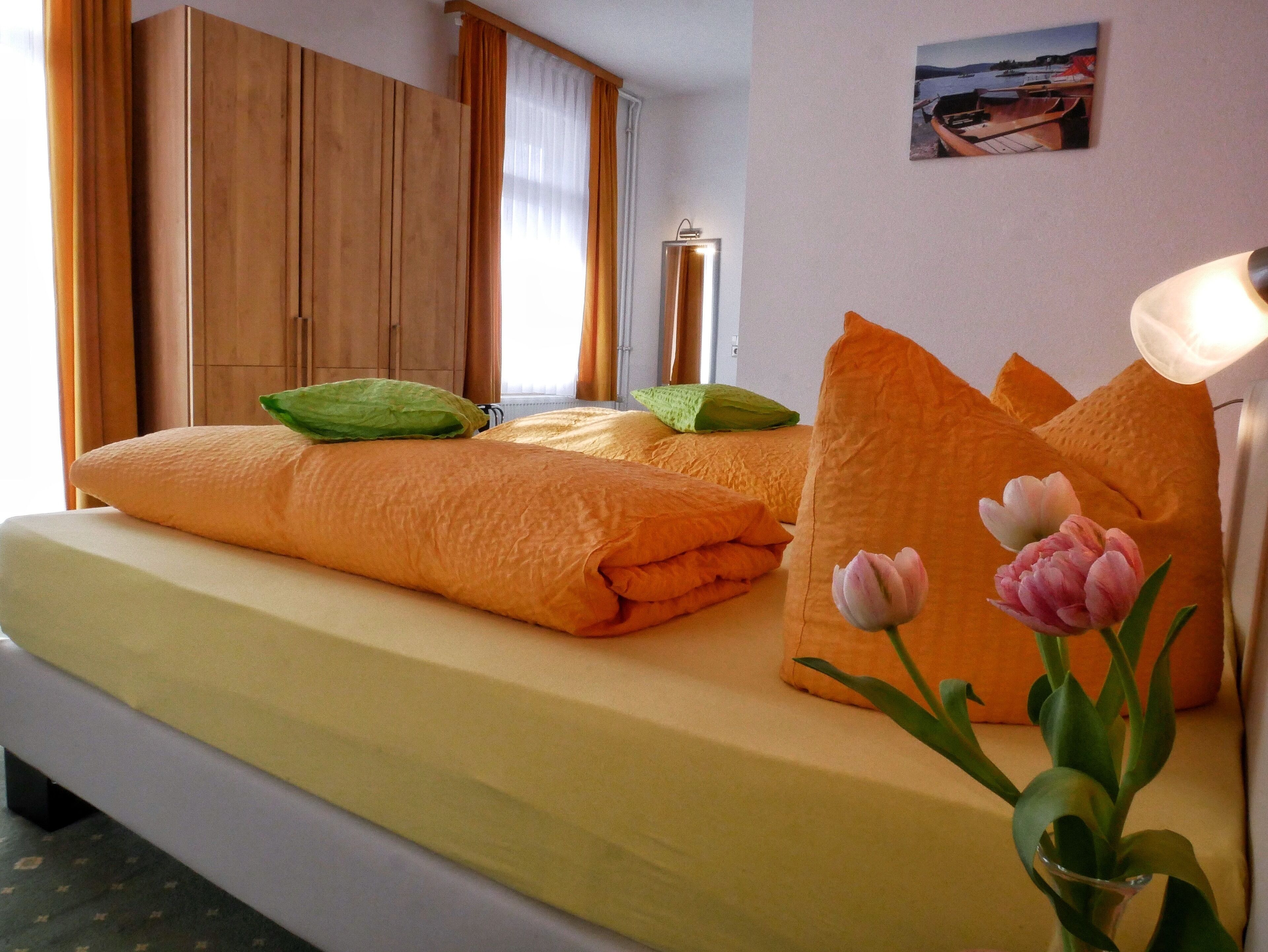 Quarto de hotel com cama, almofadas, mesa de cabeceira, abajur, quadro na parede e janela com cortinas.