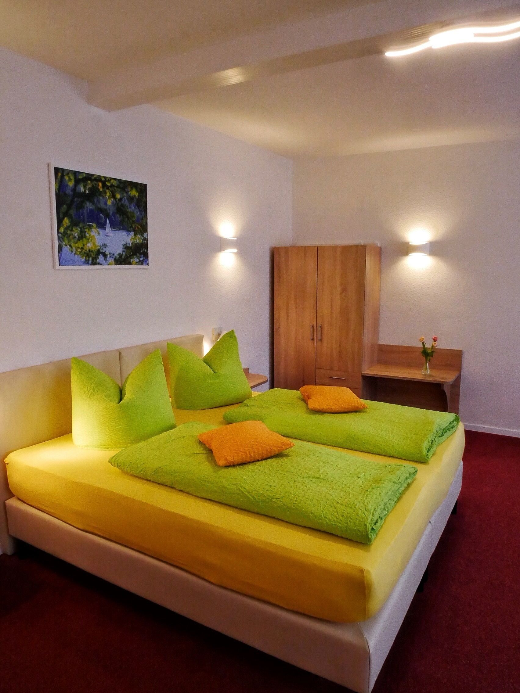 Un lit double avec des oreillers verts et orange, une armoire en bois, une petite table avec un vase et un tableau accroché au mur.
