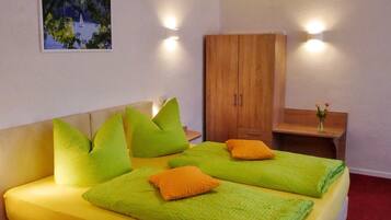 Un lit double avec des oreillers verts et orange, une armoire en bois, une petite table avec un vase et un tableau accroché au mur.