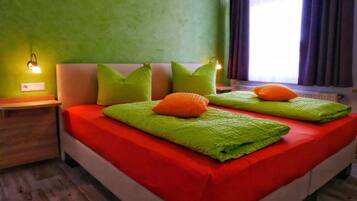 Cama doble con ropa de cama verde y naranja, una mesita de noche con lámpara y una ventana con cortinas.