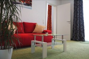 Ein Zimmer mit einem roten Sofa, einem Glastisch, einer Zimmerpflanze und einer Tür mit gemusterten Vorhängen.