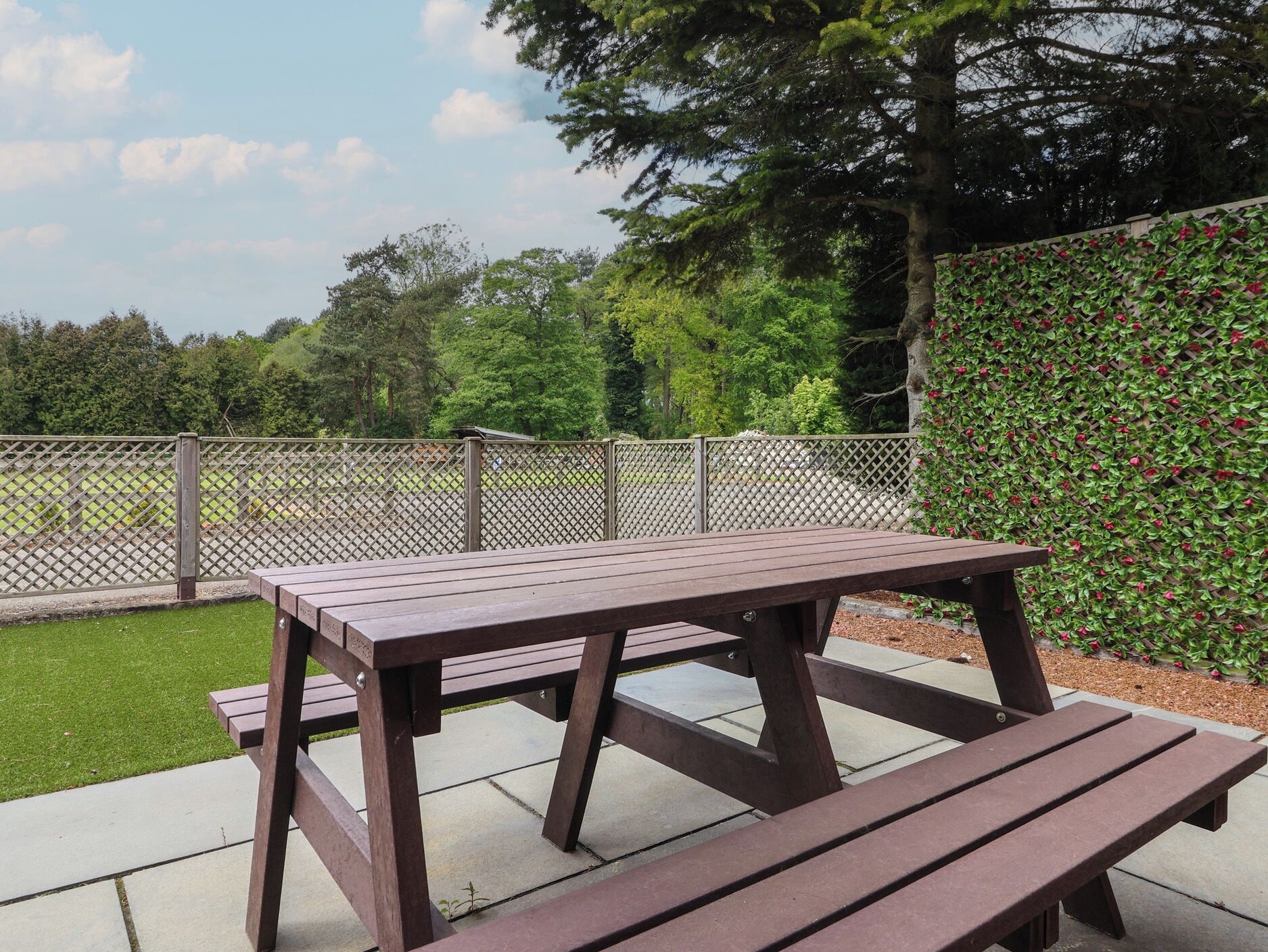 A picnic table with benches, surrounded by a wooden fence and a hedge with red flowers.