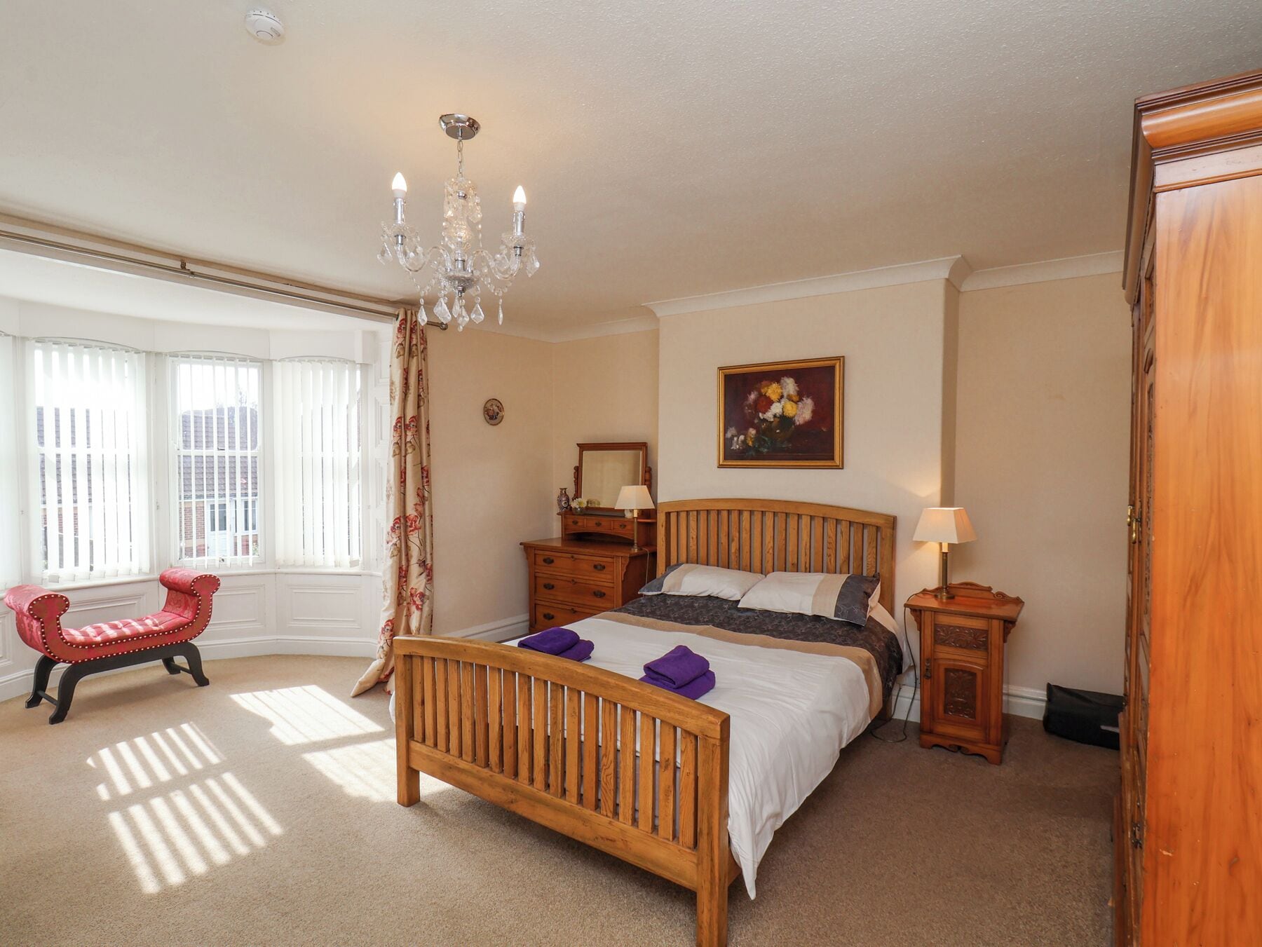 A bedroom with a wooden bed, a red armchair, a chandelier, a painting, and a window with curtains.