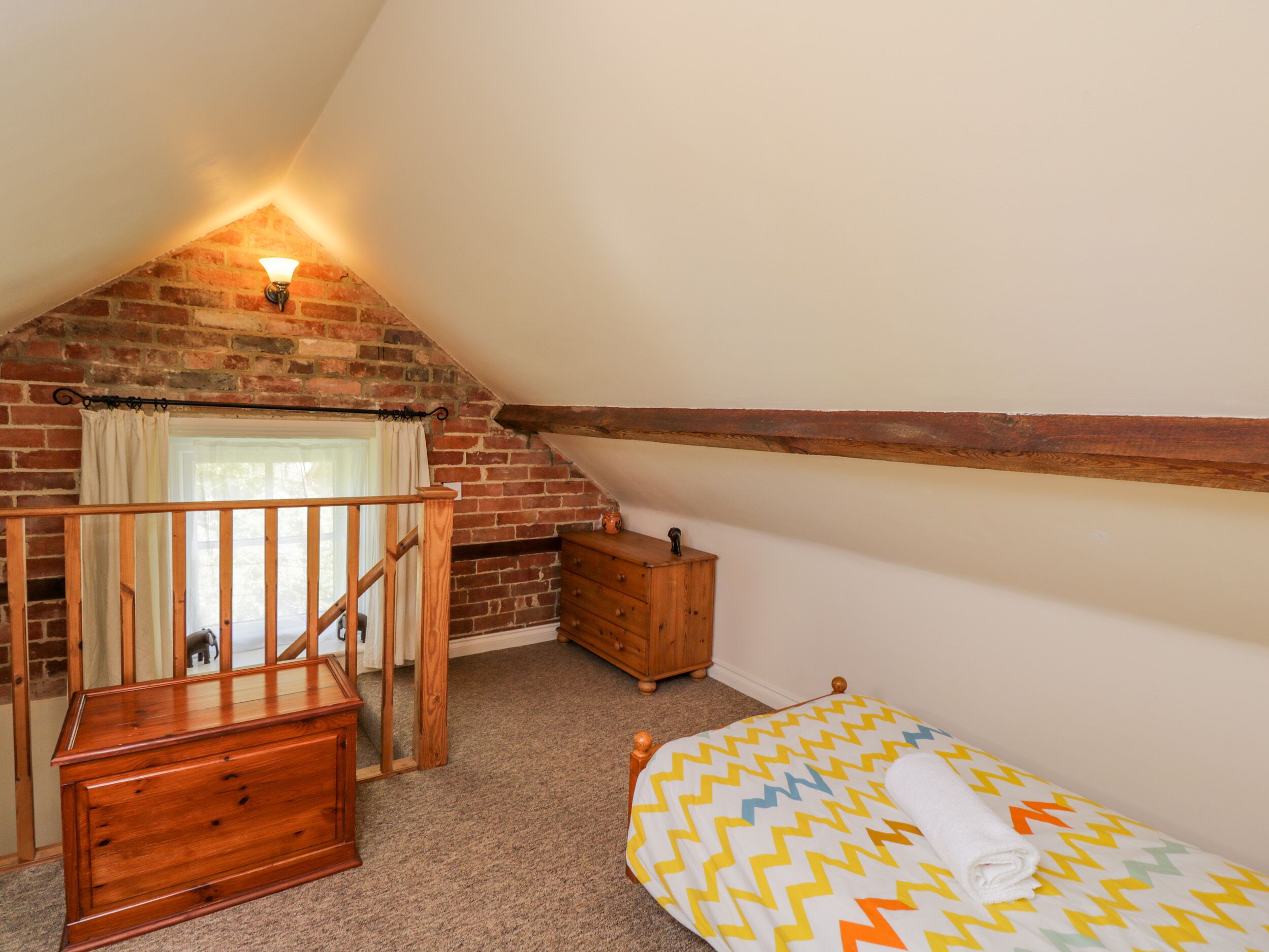 A bedroom with a sloped ceiling, a wooden dresser, a bed with a patterned blanket, and a window with curtains.
