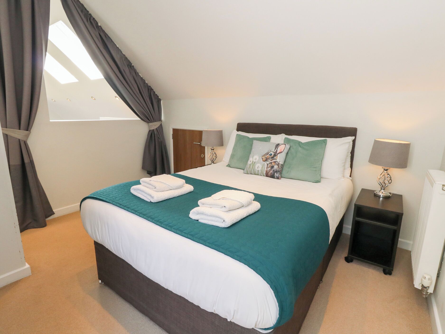 A neatly made bed with a teal bedspread and white linens, a bedside table with a lamp, and a window with curtains.
