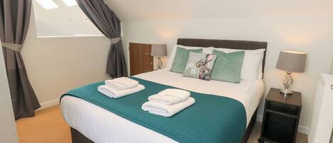 A neatly made bed with a teal bedspread and white linens, a bedside table with a lamp, and a window with curtains.