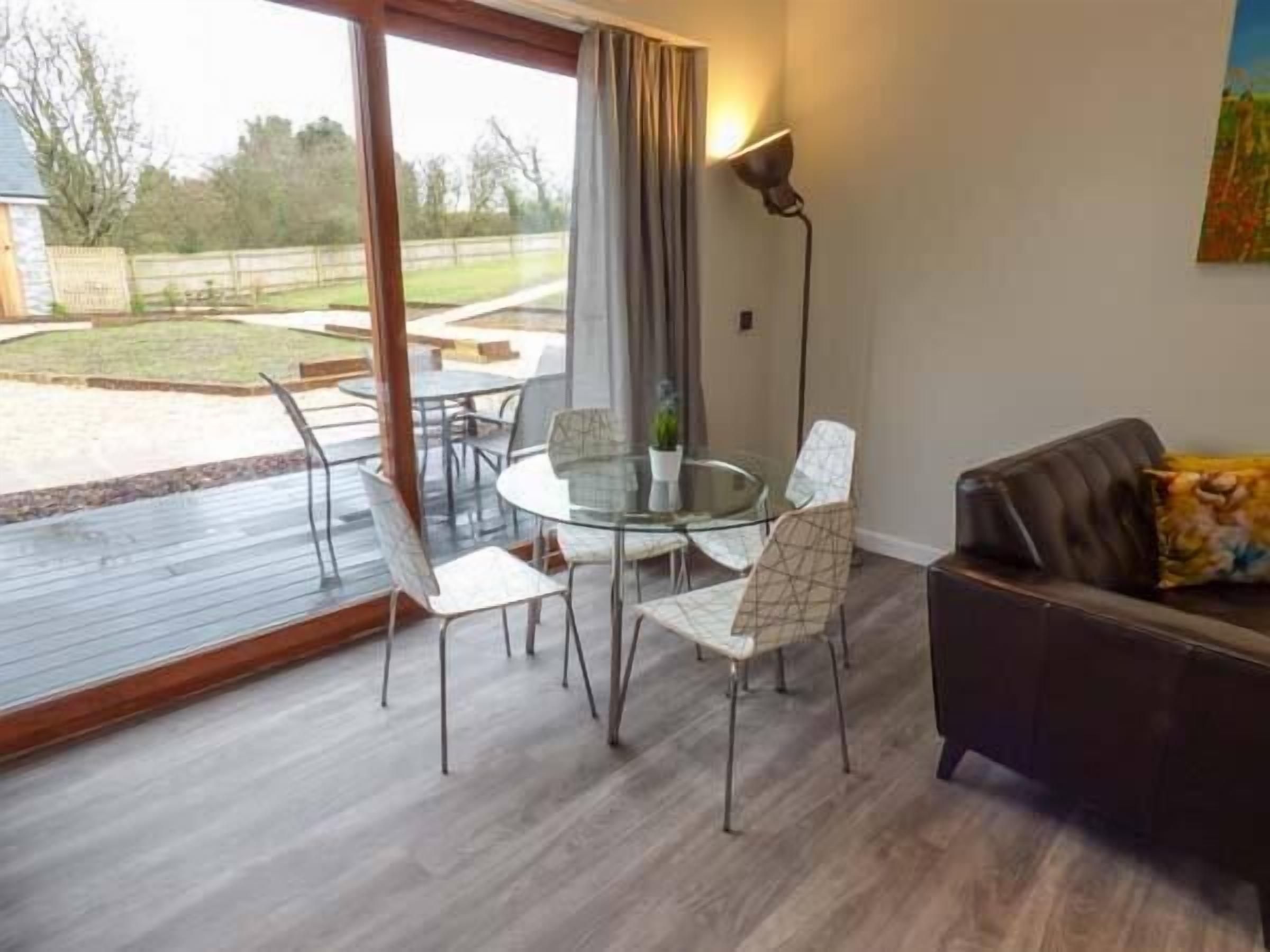 Un salon moderne avec une table et des chaises en verre, un canapé foncé et une grande fenêtre avec des rideaux.