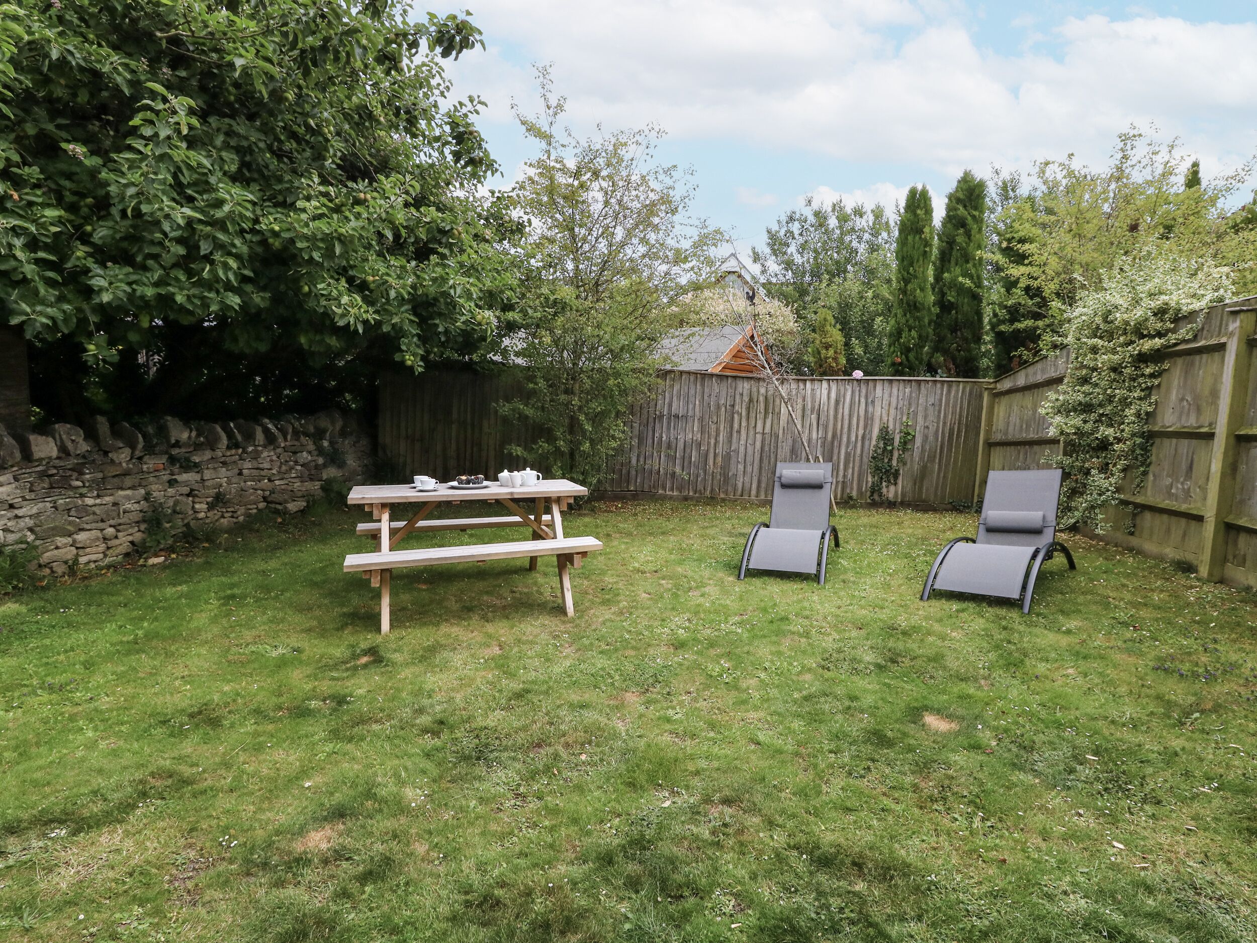 A backyard with a wooden picnic table, two lounge chairs, and a stone wall.