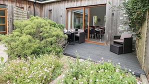 Una casa moderna con fachada de madera, un patio con mobiliario exterior y un jardín con flores rosadas.