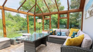 A conservatory with wicker furniture, a glass table, and a view of a lake. - Dragonfly Lodge (Dereham)