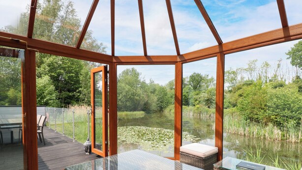 A glass-enclosed deck with a table and chairs, overlooking a pond with lily pads. - Dragonfly Lodge (Dereham)