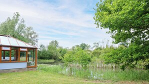 A conservatory with a glass roof, surrounded by a pond and lush greenery. - Dragonfly Lodge (Dereham)