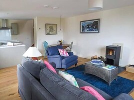 A living room with a sofa, a blue armchair, a small table with books, a fireplace, and a kitchen area in the background.