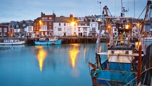 A harbor with boats docked, buildings in the background, and a crane. - Budmouth (Weymouth)